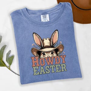 May include: A blue Comfort Colors t-shirt featuring a cartoon bunny wearing a cowboy hat. The bunny is surrounded by small flowers. The words "HOWDY EASTER" are printed in orange, green, and brown.
