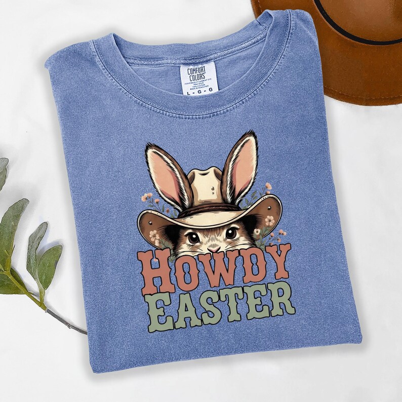 May include: A blue Comfort Colors t-shirt featuring a cartoon bunny wearing a cowboy hat. The bunny is surrounded by small flowers. The words "HOWDY EASTER" are printed in orange, green, and brown.