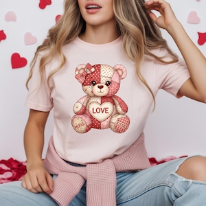 May include: A light pink t-shirt featuring a patchwork teddy bear design. The bear has a red and pink color scheme with a heart that says "LOVE". The bear also has the words "HUGS" and "KISSES" on its paws. A bow is on the bear's head.