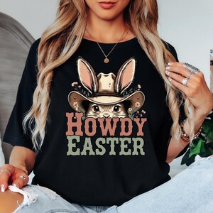 May include: Black t-shirt featuring a graphic of a bunny wearing a cowboy hat with the text "HOWDY EASTER" in pink and green. The bunny's ears and hat are tan, and the shirt is a solid black color.