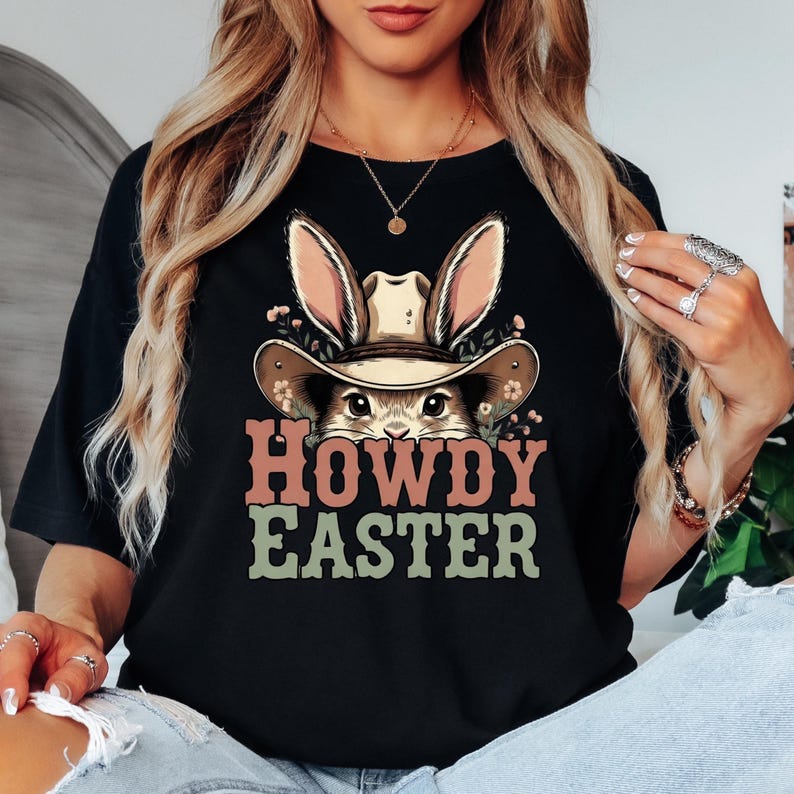 May include: Black t-shirt featuring a graphic of a bunny wearing a cowboy hat with the text "HOWDY EASTER" in pink and green. The bunny's ears and hat are tan, and the shirt is a solid black color.