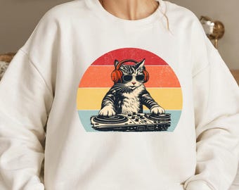 May include: White sweatshirt featuring a graphic of a cat wearing sunglasses and headphones, operating a DJ turntable. The cat is set against a retro sunset design with red, orange, yellow, and blue stripes. A fun, music-themed design.