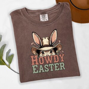 May include: Brown t-shirt with a graphic of a bunny wearing a cowboy hat and the words "HOWDY EASTER" in pink and green. The shirt is folded and has a Comfort Colors tag.