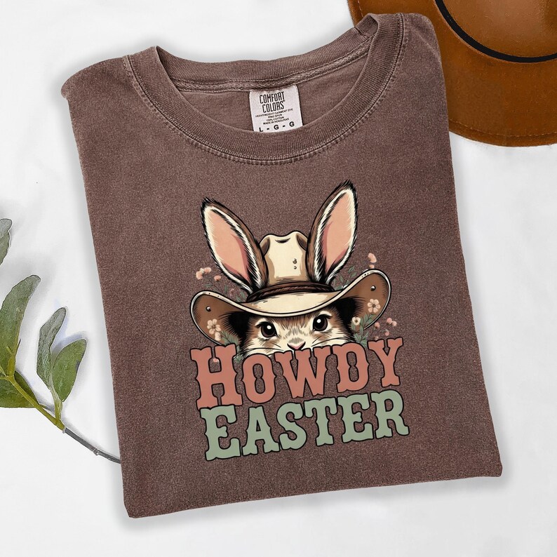 May include: Brown t-shirt with a graphic of a bunny wearing a cowboy hat and the words "HOWDY EASTER" in pink and green. The shirt is folded and has a Comfort Colors tag.