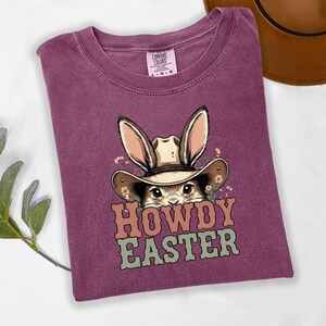 May include: A purple t-shirt with a graphic of a bunny wearing a cowboy hat. The bunny is surrounded by flowers, and the text "HOWDY EASTER" is printed below the bunny in orange and green letters. The shirt is folded.
