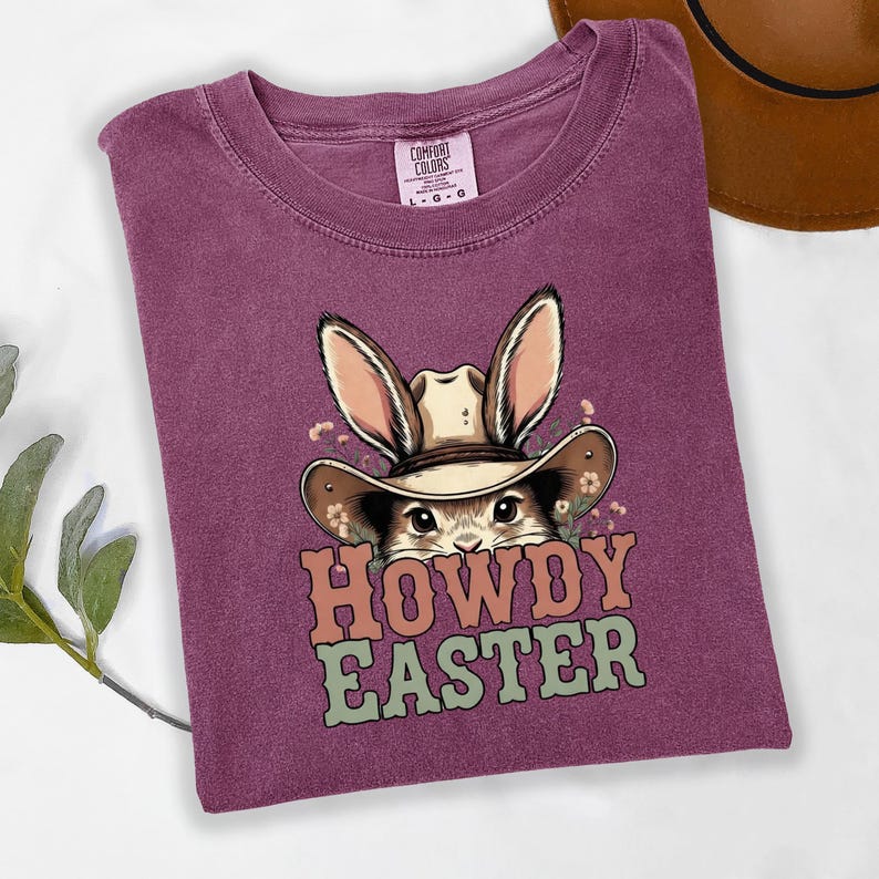 May include: A purple t-shirt with a graphic of a bunny wearing a cowboy hat. The bunny is surrounded by flowers, and the text "HOWDY EASTER" is printed below the bunny in orange and green letters. The shirt is folded.