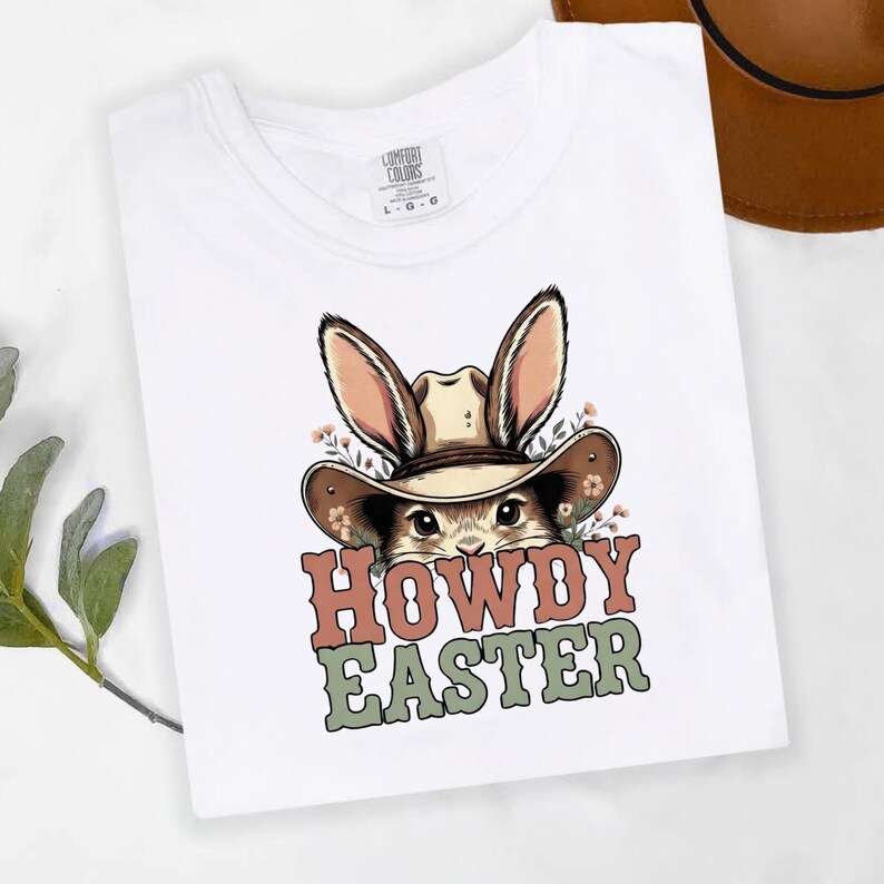 May include: White t-shirt featuring a cartoon bunny wearing a cowboy hat, with the text "HOWDY EASTER" in a western-style font. The bunny has long ears and is surrounded by small flowers. The shirt is made of a soft material.
