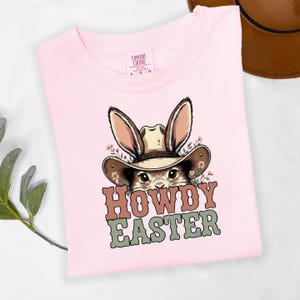 May include: A light pink t-shirt featuring a cartoon bunny wearing a cowboy hat. The bunny is surrounded by flowers and the text "HOWDY EASTER" is printed in brown and green. The shirt has a Comfort Colors tag.