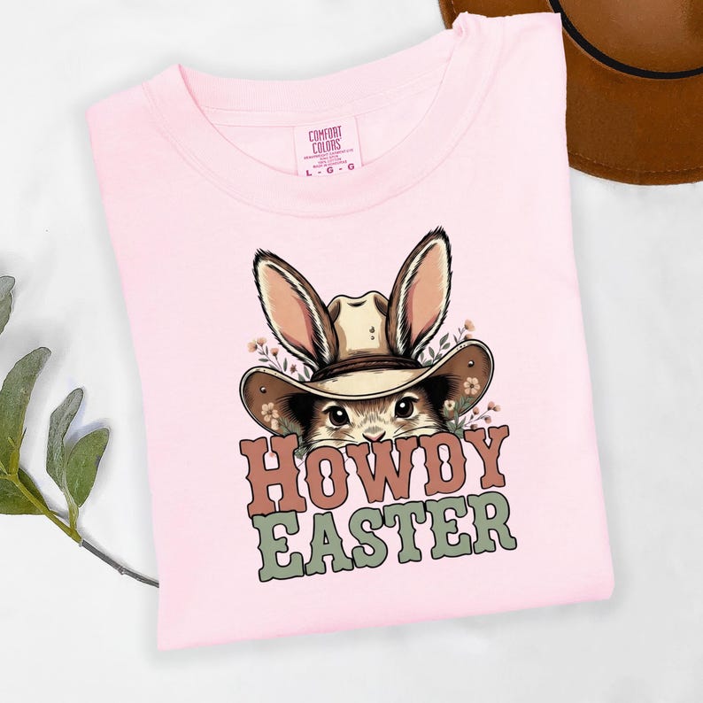May include: A light pink t-shirt featuring a cartoon bunny wearing a cowboy hat. The bunny is surrounded by flowers and the text "HOWDY EASTER" is printed in brown and green. The shirt has a Comfort Colors tag.