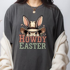 May include: Dark gray t-shirt featuring a cartoon bunny wearing a cowboy hat with the text "HOWDY EASTER" in orange and green. The bunny has long ears and is surrounded by small flowers. A casual, festive design.