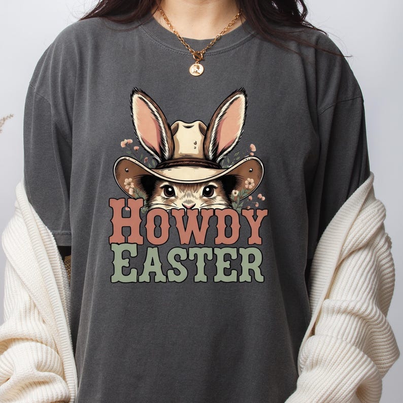 May include: Dark gray t-shirt featuring a cartoon bunny wearing a cowboy hat with the text "HOWDY EASTER" in orange and green. The bunny has long ears and is surrounded by small flowers. A casual, festive design.