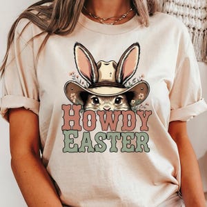 May include: A beige t-shirt featuring a graphic of a bunny wearing a cowboy hat with bunny ears. The text "HOWDY EASTER" is in pink and green. The shirt has rolled-up sleeves.