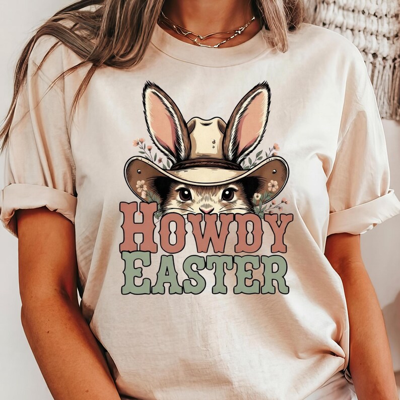 May include: A beige t-shirt featuring a graphic of a bunny wearing a cowboy hat with bunny ears. The text "HOWDY EASTER" is in pink and green. The shirt has rolled-up sleeves.