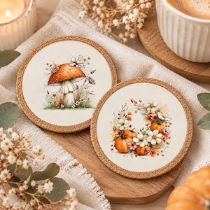 Könnte beinhalten: Zwei runde Untersetzer mit Korkrückseite und weißer Oberfläche. Ein Untersetzer zeigt eine Illustration eines großen orangefarbenen Pilzes, umgeben von kleinen weißen Blumen. Der andere zeigt einen Kranz aus orangefarbenen Kürbissen und weißen Blumen.