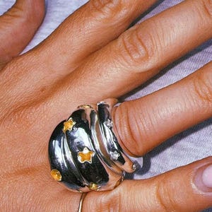 Pu&ograve; includere: Un ampio anello in argento lucido caratterizzato da un design a spirale a pi&ugrave; strati. La superficie &egrave; decorata con diverse piccole pietre preziose gialle a forma di stella e pietre trasparenti dal taglio rotondo.