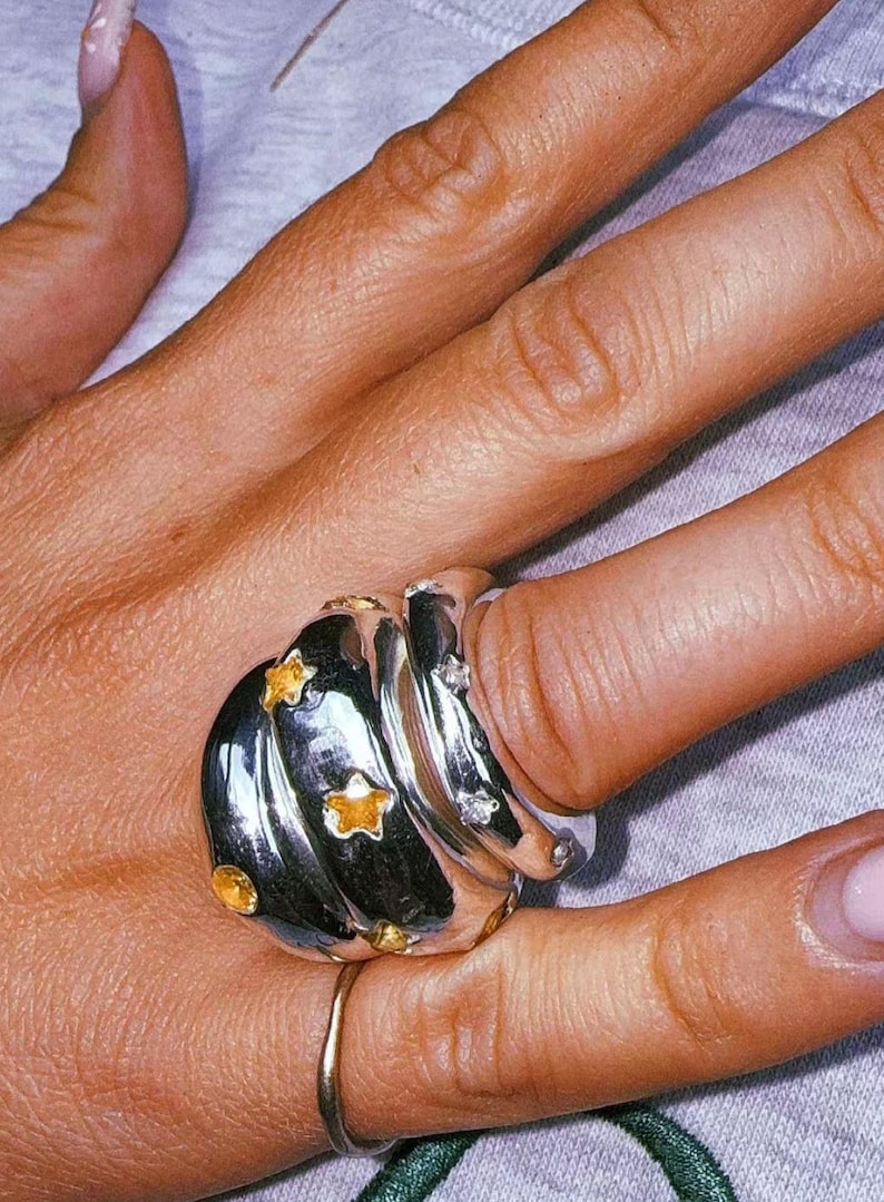 Pu&ograve; includere: Un ampio anello in argento lucido caratterizzato da un design a spirale a pi&ugrave; strati. La superficie &egrave; decorata con diverse piccole pietre preziose gialle a forma di stella e pietre trasparenti dal taglio rotondo.
