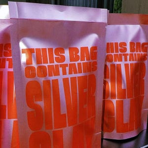 Könnte beinhalten: Drei hellrosa Standbeutel mit kräftigem orangefarbenem Text, der THIS BAG CONTAINS SILVER CLAY lautet. Die Beutel haben eine matte Oberfläche und eine heißversiegelte Oberkante und stehen in einer Reihe, wobei der Text deutlich sichtbar ist.