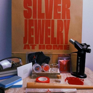 Pu&ograve; includere: Un kit per la creazione di gioielli in argento con un sacchetto di carta marrone stampato con il testo THIS BAG CONTAINS EVERYTHING TO MAKE YOUR OWN SILVER JEWELRY AT HOME, un cannello nero, occhiali protettivi, utensili in legno e metallo.