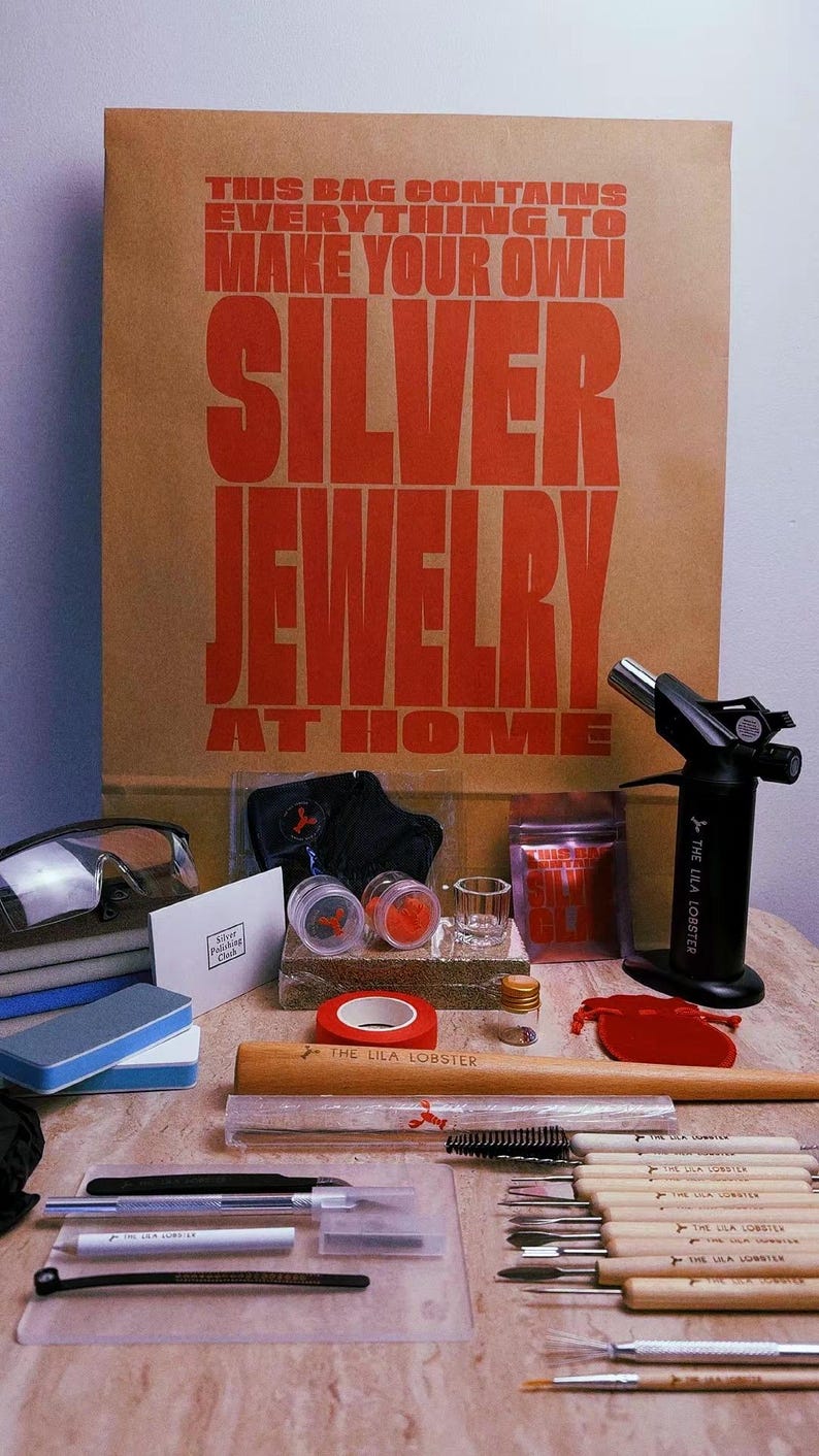 Pu&ograve; includere: Un kit per la creazione di gioielli in argento con un sacchetto di carta marrone stampato con il testo THIS BAG CONTAINS EVERYTHING TO MAKE YOUR OWN SILVER JEWELRY AT HOME, un cannello nero, occhiali protettivi, utensili in legno e metallo.
