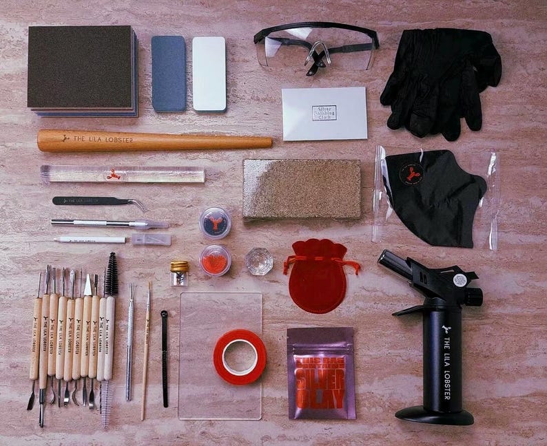 Pu&ograve; includere: Un kit per la lavorazione dell'argilla metallica che include un cannello a butano nero, occhiali protettivi, guanti neri, un mandrino in legno per anelli, vari strumenti di scultura, blocchi abrasivi, un sacchetto di velluto rosso, nastro rosso e argilla d'argento.