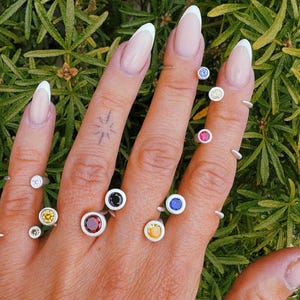 Pu&ograve; includere: Una collezione di anelli in argento con piccole pietre preziose rotonde incastonate a lunetta in vari colori, tra cui rosso, blu, giallo e bianco. Gli anelli sono indossati su pi&ugrave; dita, mostrando una variet&agrave; di pietre lucide e vivaci.