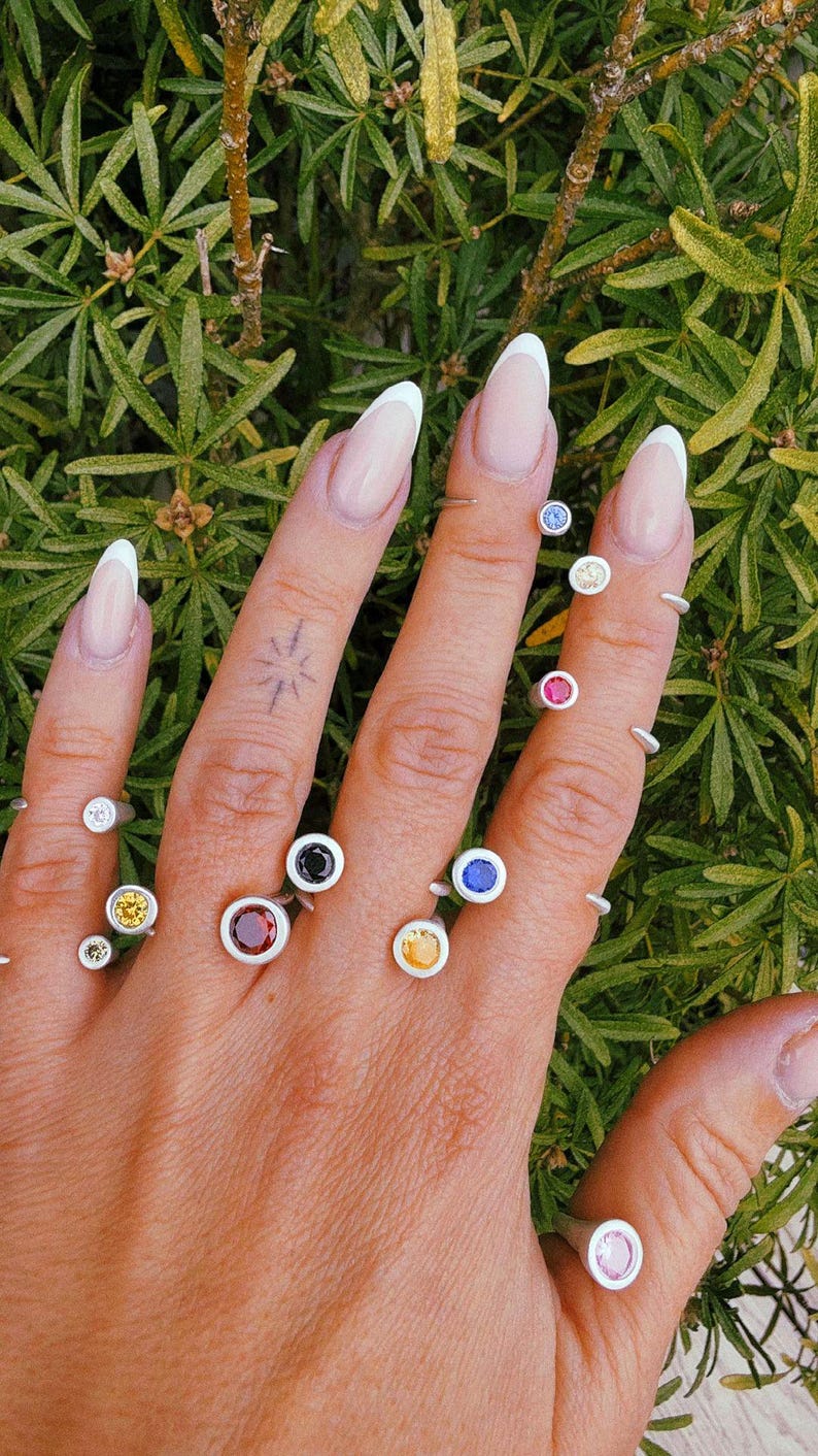 Pu&ograve; includere: Una collezione di anelli in argento con piccole pietre preziose rotonde incastonate a lunetta in vari colori, tra cui rosso, blu, giallo e bianco. Gli anelli sono indossati su pi&ugrave; dita, mostrando una variet&agrave; di pietre lucide e vivaci.