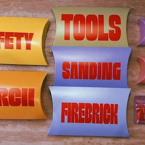 Pu&ograve; includere: Una collezione di scatole a cuscino di vari colori con etichette in testo rosso tra cui SAFETY, TORCH, TOOLS, SANDING, FIREBRICK, SHINE e GEMS. Sono inclusi anche una piccola bustina argentata con etichetta SILVER CLAY e un sacchetto rosso scuro.