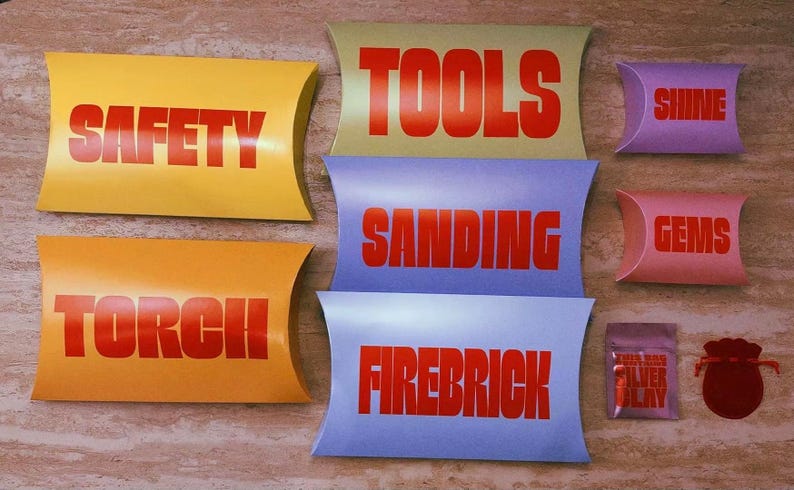 Pu&ograve; includere: Una collezione di scatole a cuscino di vari colori con etichette in testo rosso tra cui SAFETY, TORCH, TOOLS, SANDING, FIREBRICK, SHINE e GEMS. Sono inclusi anche una piccola bustina argentata con etichetta SILVER CLAY e un sacchetto rosso scuro.