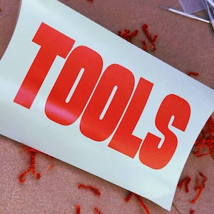 Puede incluir: Un letrero rectangular blanco con la palabra TOOLS impresa en letras mayúsculas rojas brillantes y negritas. El letrero está rodeado de trozos de papel rojo esparcidos y varios suministros de arte, incluyendo pinceles finos y un recipiente.
