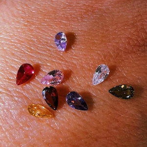 May include: A collection of eight small, pear-shaped faceted gemstones in various colors, including red, yellow, purple, pink, dark brown, olive green, and clear white, arranged in a scattered pattern on a textured skin surface.