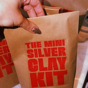 以下が含まれることがあります： 前面に太い赤色の大文字で「THE MINI SILVER CLAY KIT」と印刷された茶色の紙袋。この袋は標準的なクラフト紙素材で作られており、上端が折り返された長方形の形状をしています。