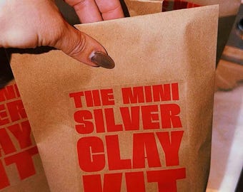 以下が含まれることがあります： 前面に太い赤色の大文字で「THE MINI SILVER CLAY KIT」と印刷された茶色の紙袋。この袋は標準的なクラフト紙素材で作られており、上端が折り返された長方形の形状をしています。