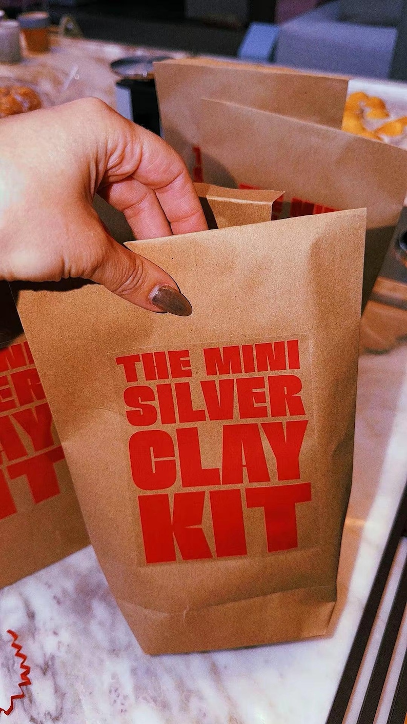 May include: A brown paper bag with the text THE MINI SILVER CLAY KIT printed in bold red capital letters on the front. The bag is made of a standard kraft paper material and features a rectangular shape with a folded top edge for closure.