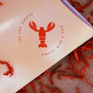 Puede incluir: Una caja de cartón de color beige claro con un gráfico de langosta roja en el centro. El texto que rodea a la langosta dice THE LILA LOBSTER JEWELRY WITH A TALE. La caja está rodeada de papel de relleno rojo cortado en tiras sobre una superficie.