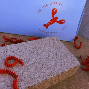 Op de afbeelding: Een lichtblauwe papieren zak met een rode kreeftafbeelding en de tekst THE LILA LOBSTER JEWELRY WITH A TALE. De zak staat achter een rechthoekig grijs stenen blok, met verspreide rode stroken gekreukt papier rondom de voorwerpen.