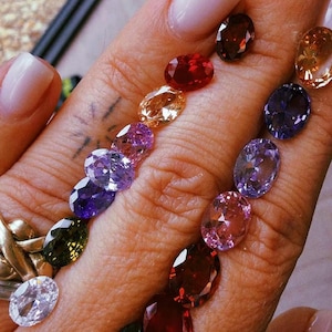 May include: A collection of oval-cut faceted gemstones in a variety of colors including red, purple, pink, green, orange, and clear, arranged in two rows. The stones exhibit high clarity and reflective surfaces, showcasing a spectrum of vibrant hues.