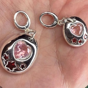 Pu&ograve; includere: Due ciondoli color argento con chiusure a moschettone. Ogni ciondolo presenta una base metallica liscia e arrotondata con incastonata una gemma rosa a forma di cuore e tre gemme rosse pi&ugrave; piccole a forma di stella.
