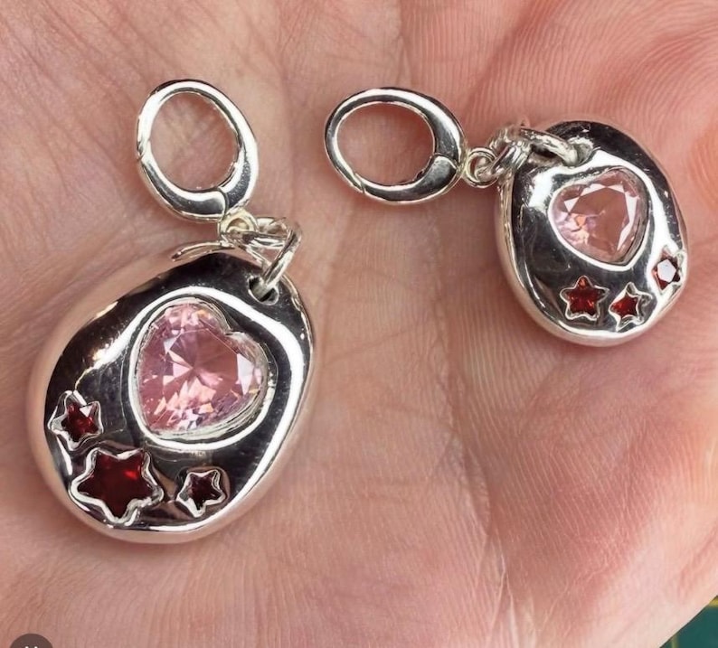 Pu&ograve; includere: Due ciondoli color argento con chiusure a moschettone. Ogni ciondolo presenta una base metallica liscia e arrotondata con incastonata una gemma rosa a forma di cuore e tre gemme rosse pi&ugrave; piccole a forma di stella.