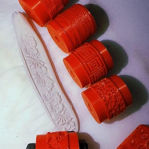 Peut inclure: Une collection de rouleaux à argile cylindriques orange présentant divers motifs en relief, notamment des motifs de vannerie, linéaires, géométriques et floraux. Un rouleau est fixé à un manche noir et une bande d'argile blanche montre une empreinte florale.