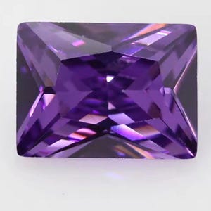 May include: A rectangular-cut gemstone with a deep purple hue and a faceted surface that creates geometric light reflections. The stone features sharp, clean edges and a symmetrical arrangement of triangular and trapezoidal facets across its top.