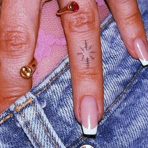 Pu&ograve; includere: Due anelli d'oro con piccole pietre circolari, una rossa e una gialla, indossati sulle dita. Un dito presenta un piccolo tatuaggio a forma di stella con inchiostro nero. Le dita sono appoggiate su tessuto denim blu con bottone metallico.