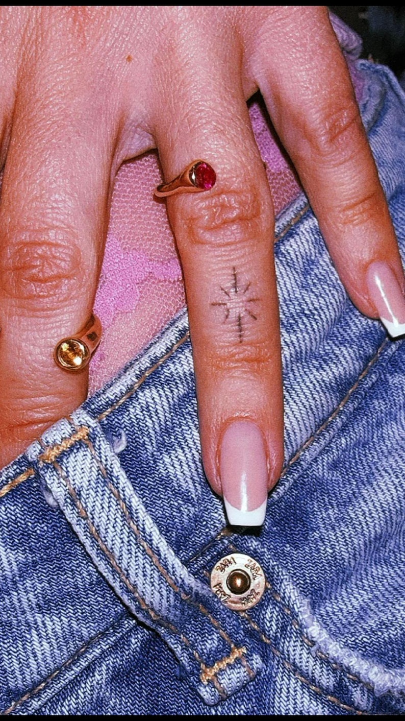 Pu&ograve; includere: Due anelli d'oro con piccole pietre circolari, una rossa e una gialla, indossati sulle dita. Un dito presenta un piccolo tatuaggio a forma di stella con inchiostro nero. Le dita sono appoggiate su tessuto denim blu con bottone metallico.