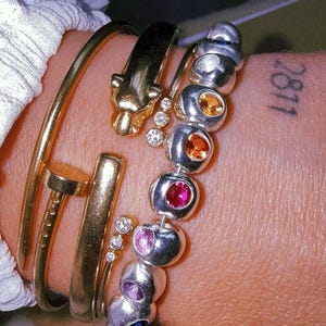 Pu&ograve; includere: Un bracciale rigido dorato a forma di chiodo, un bracciale dorato con testa di pantera e tre piccoli diamanti, e un bracciale di perline argentate con una fila di pietre preziose multicolori arancioni, rosse, rosa, viola e blu incastonate.