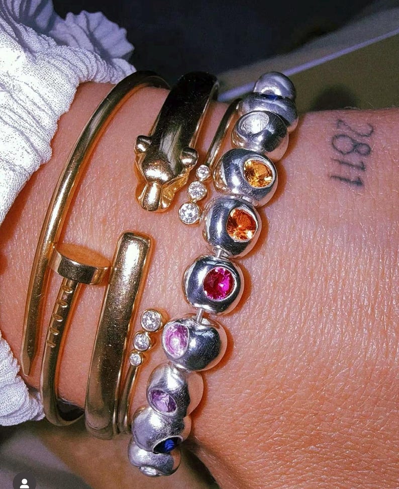 Pu&ograve; includere: Un bracciale rigido dorato a forma di chiodo, un bracciale dorato con testa di pantera e tre piccoli diamanti, e un bracciale di perline argentate con una fila di pietre preziose multicolori arancioni, rosse, rosa, viola e blu incastonate.