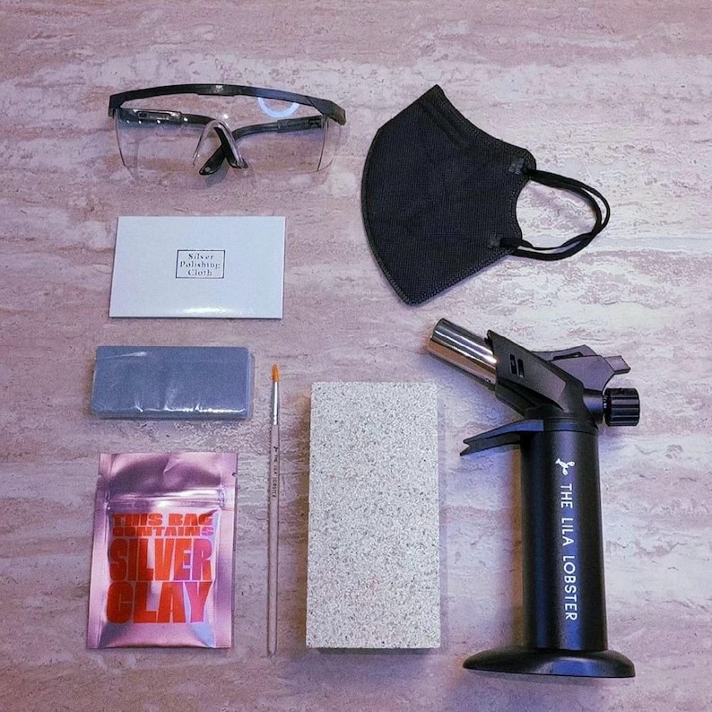 May include: A metal torch with the text THE LILA LOBSTER, a black face mask, clear safety glasses, a silver polishing cloth, a blue rectangular block, a small brush, a grey textured block, and a pink pouch labeled SILVER CLAY.