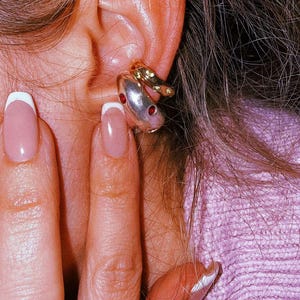 Pu&ograve; includere: Due ear cuff sovrapposti indossati sulla cartilagine. Il cerchietto superiore &egrave; una fascia sottile color oro con piccole pietre verdi incastonate. Il cerchietto inferiore &egrave; una fascia pi&ugrave; spessa e liscia color argento con due pietre rosse circolari.