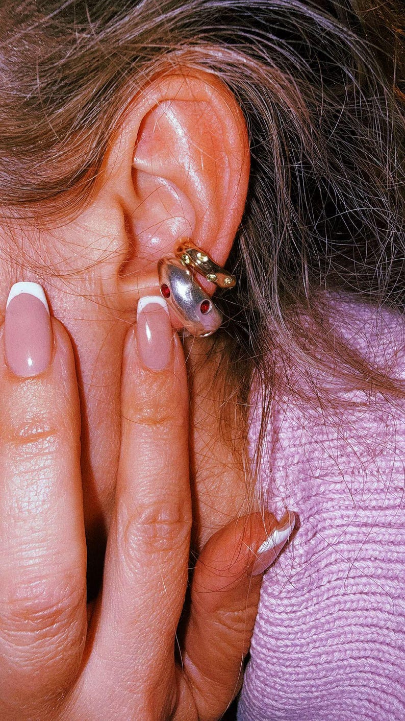 Pu&ograve; includere: Due ear cuff sovrapposti indossati sulla cartilagine. Il cerchietto superiore &egrave; una fascia sottile color oro con piccole pietre verdi incastonate. Il cerchietto inferiore &egrave; una fascia pi&ugrave; spessa e liscia color argento con due pietre rosse circolari.