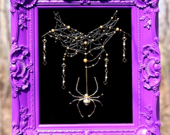 May include: A framed artwork featuring a spider web and spider design. The frame is a vibrant purple with ornate detailing. The spider web is crafted with delicate gold beads and the spider has a detailed body. The background is black.