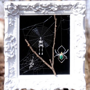 May include: A white ornate framed art piece with a black background. A spider web, skeleton, snake, and spider with a green gem are displayed. The art is mounted on a wooden post in a natural outdoor setting.