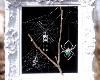 May include: A white ornate framed art piece with a black background. A spider web, skeleton, snake, and spider with a green gem are displayed. The art is mounted on a wooden post in a natural outdoor setting.